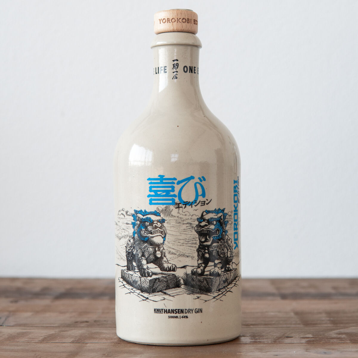 Eine Flasche Knut Hansen Sonderedition 2024 YOROKOBI steht auf einem hölzernen Tisch. Die Flasche ist größtenteils weiß mit blauen und schwarzen Designelementen, die asiatische Kunst darstellen.